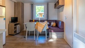 A compact living space with a dining table, a sofa, a TV, and a window with a view of greenery. - Cozy mobile home with terrace in Rivedoux-Plage for 4 ppl. (Rivedoux-Plage)