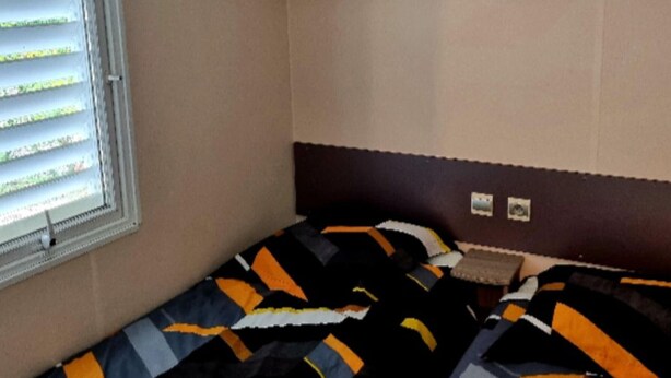A single bed with a patterned bedspread, a window with blinds, and a wall-mounted shelf. - Family mobile home with terrace, 3 bedrooms for 7 people (Nibelle)
