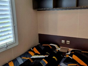 Een enkel bed met een bedovertrek met patroon, een raam met jaloezieën en een wandplank.