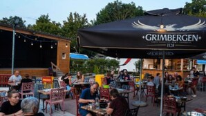 Un área de comedor al aire libre con mesas y sillas, un paraguas grande con la marca 'GRIMBERGEN' y un edificio de ladrillo al fondo.