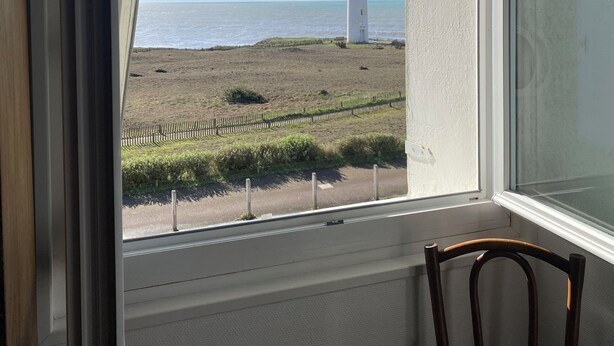 Uitzicht vanuit een kamer met een groot rondboogvenster op een grasrijk gebied met een vuurtoren.