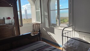 Een slaapkamer met een bed, een stoel, een dressoir en twee ramen met uitzicht op het strand.
