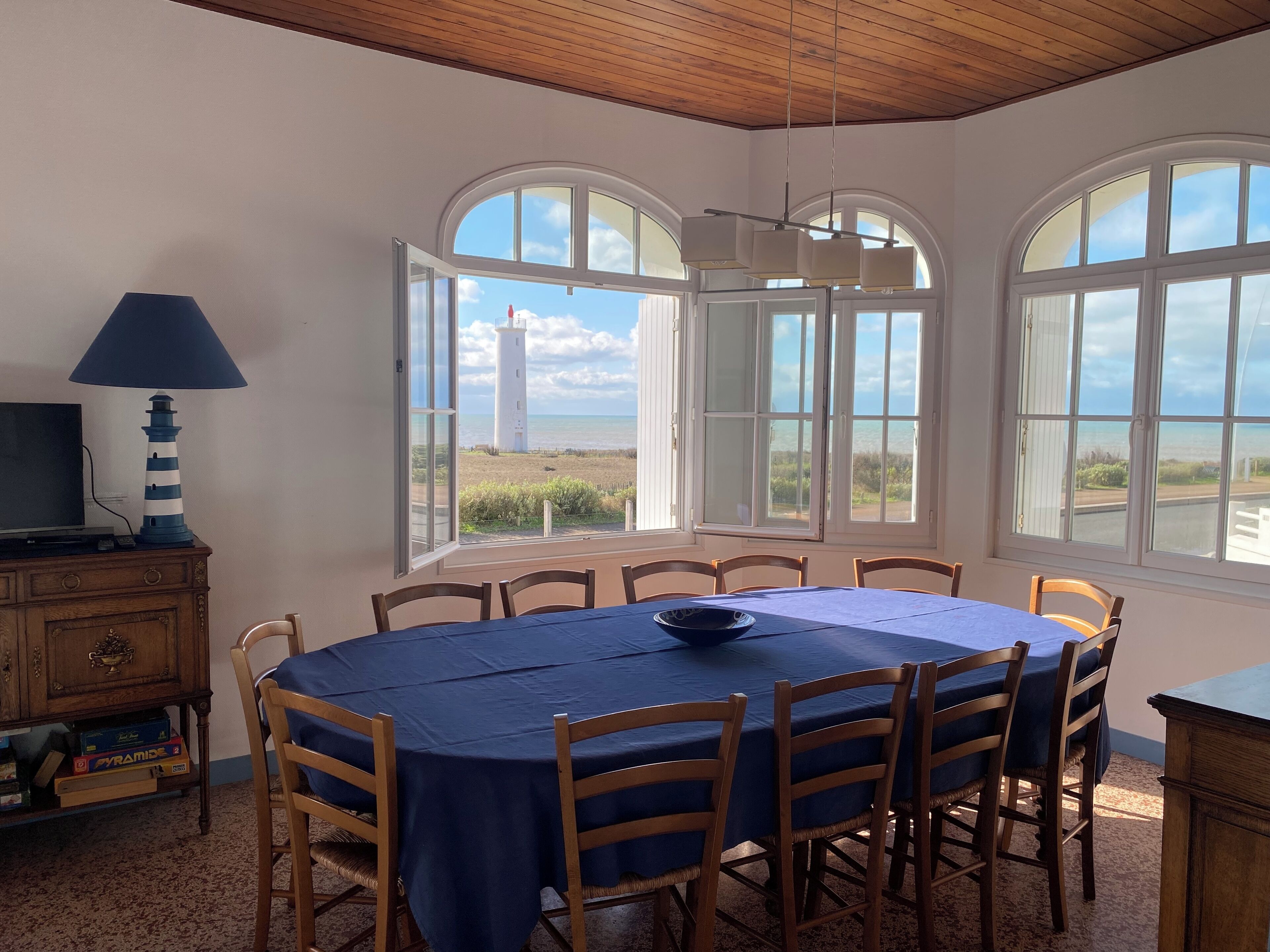 Un espace repas comprenant une table ronde, des chaises et une vue sur la plage et le phare, grâce à de grandes fenêtres.