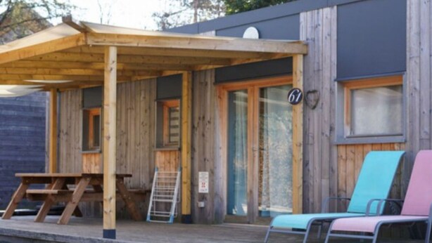 En stuga byggd av trä med en inglasad veranda, ett trädäck och två färgglada loungestolar.