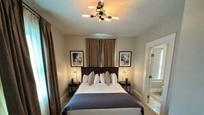 Une chambre d’hôtel bien rangée, avec un grand lit, deux tables de chevet avec des lampes, un ventilateur de plafond et deux tableaux encadrés accrochés au mur.