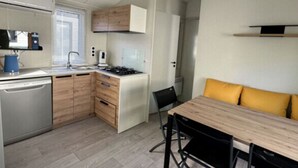 Een kleine keuken met een vaatwasser, fornuis en houten kastjes. Er is een eethoek met een tafel en stoelen.