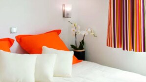 Una cama con ropa de cama blanca y naranja, una orquídea en maceta y una cortina a rayas.