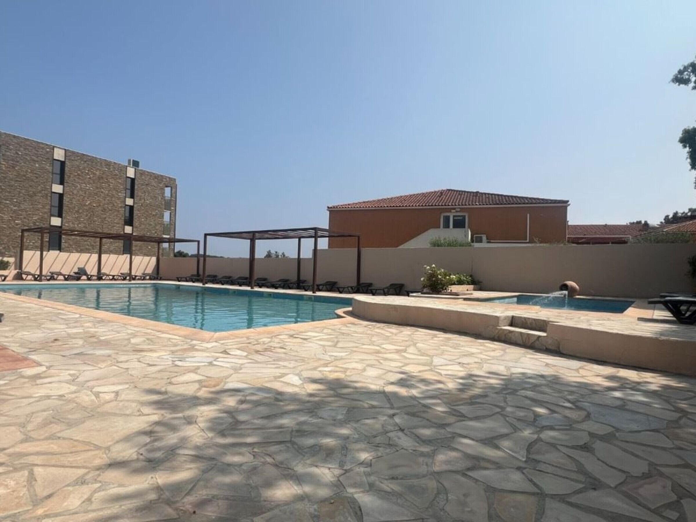 A swimming pool with lounge chairs and a pergola, surrounded by stone tiles and buildings.