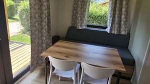 A dining area with a wooden table and two white chairs, a black sofa, and a window with patterned curtains. - Mobile Home with Terrace in Charlieu, 6 Guests (Charlieu)