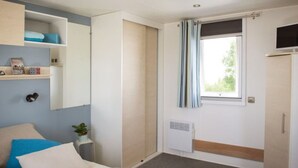 Een kleine kamer met een bed, een kledingkast, een plank met boeken, een radiator en een raam met gordijnen.