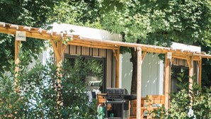 A wooden pergola with a grill and a chair in a garden setting.