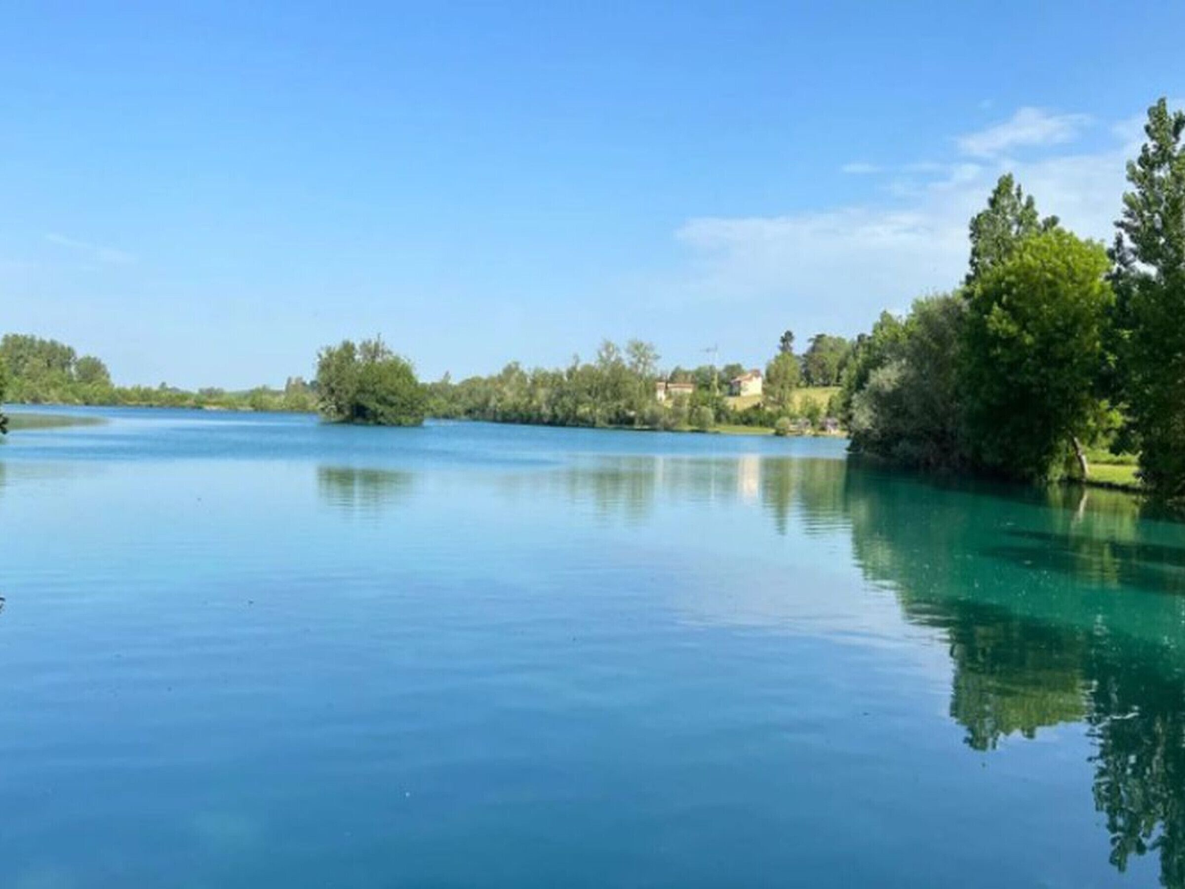 A calm river with clear water reflecting the surrounding greenery and a few buildings in the distance.