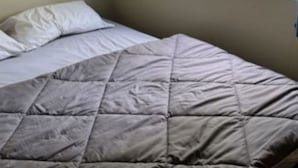 Uma cama com colcha cinza, travesseiros brancos e janela com vista para o verde.
