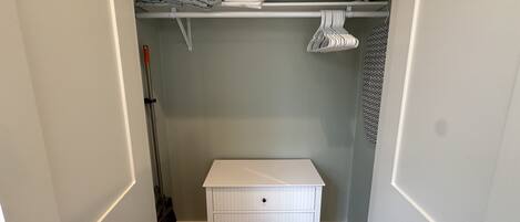 A small, neatly organized closet with a white dresser, shelves for folded clothes, and hanging garments.