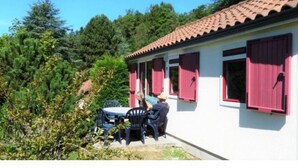 Ein weißes Gebäude mit rotem Ziegeldach und rosa Fensterläden, umgeben von Grünpflanzen.