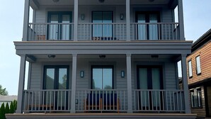 Un edificio residencial de dos pisos con un balcón en cada planta, ventanas amplias y una terraza de madera al frente.