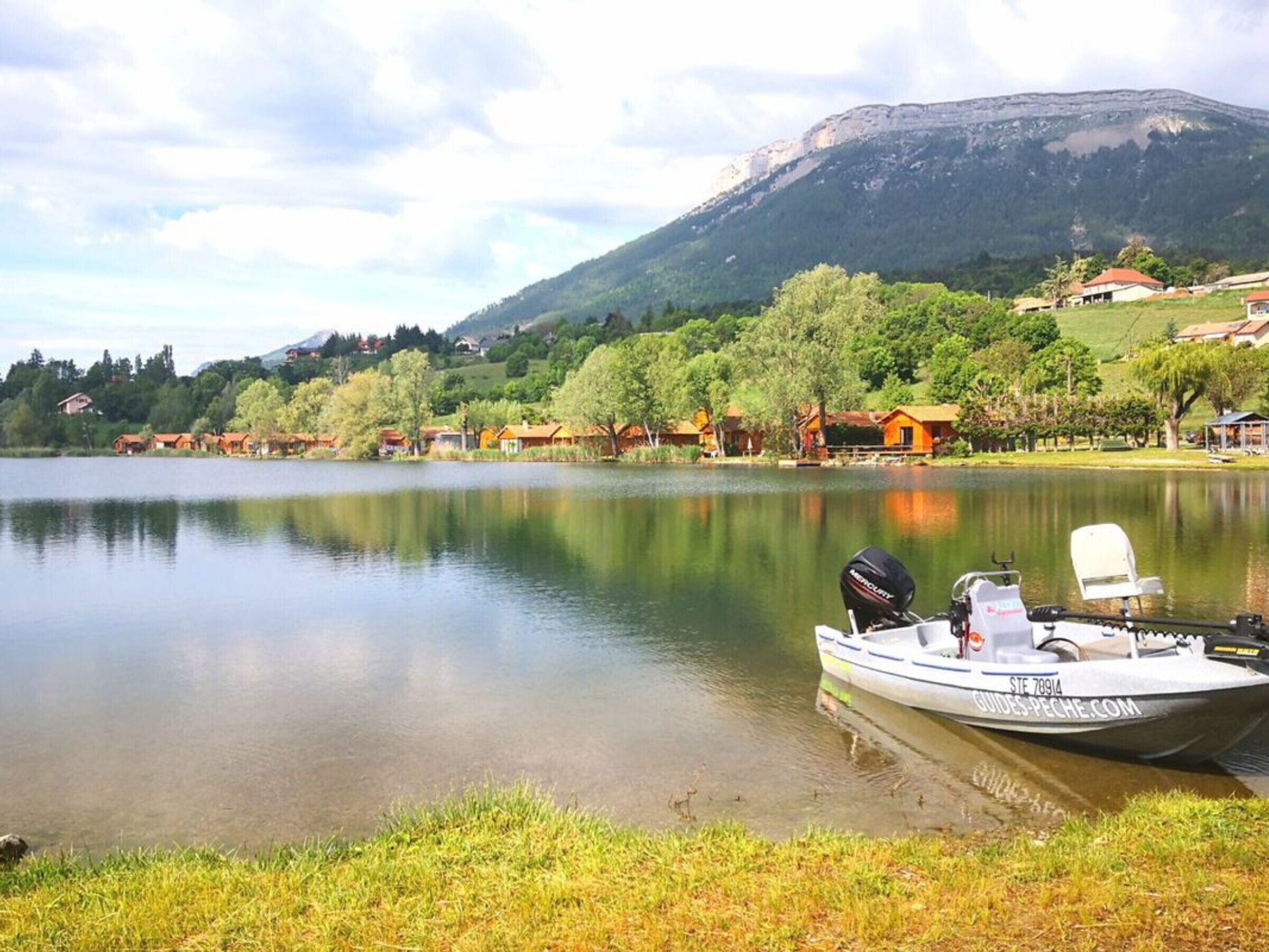 A calm lake with a boat docked on the shore, surrounded by houses and a mountain in the background.