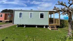 Ein eingeschossiges Mobilheim mit Veranda und zwei Fenstern, gelegen auf einer grünen Wiese unter blauem Himmel.