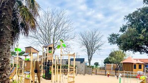 Ein Spielplatz mit verschiedenen Spielgeräten, umgeben von einem Holzzaun und Palmen.