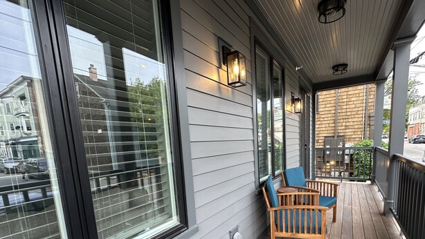 A covered porch with two wooden chairs and a table, large windows, and exterior lighting fixtures. - Brand New! 2 Bed W/ Private Deck 2 Bedroom Apts (Newport)