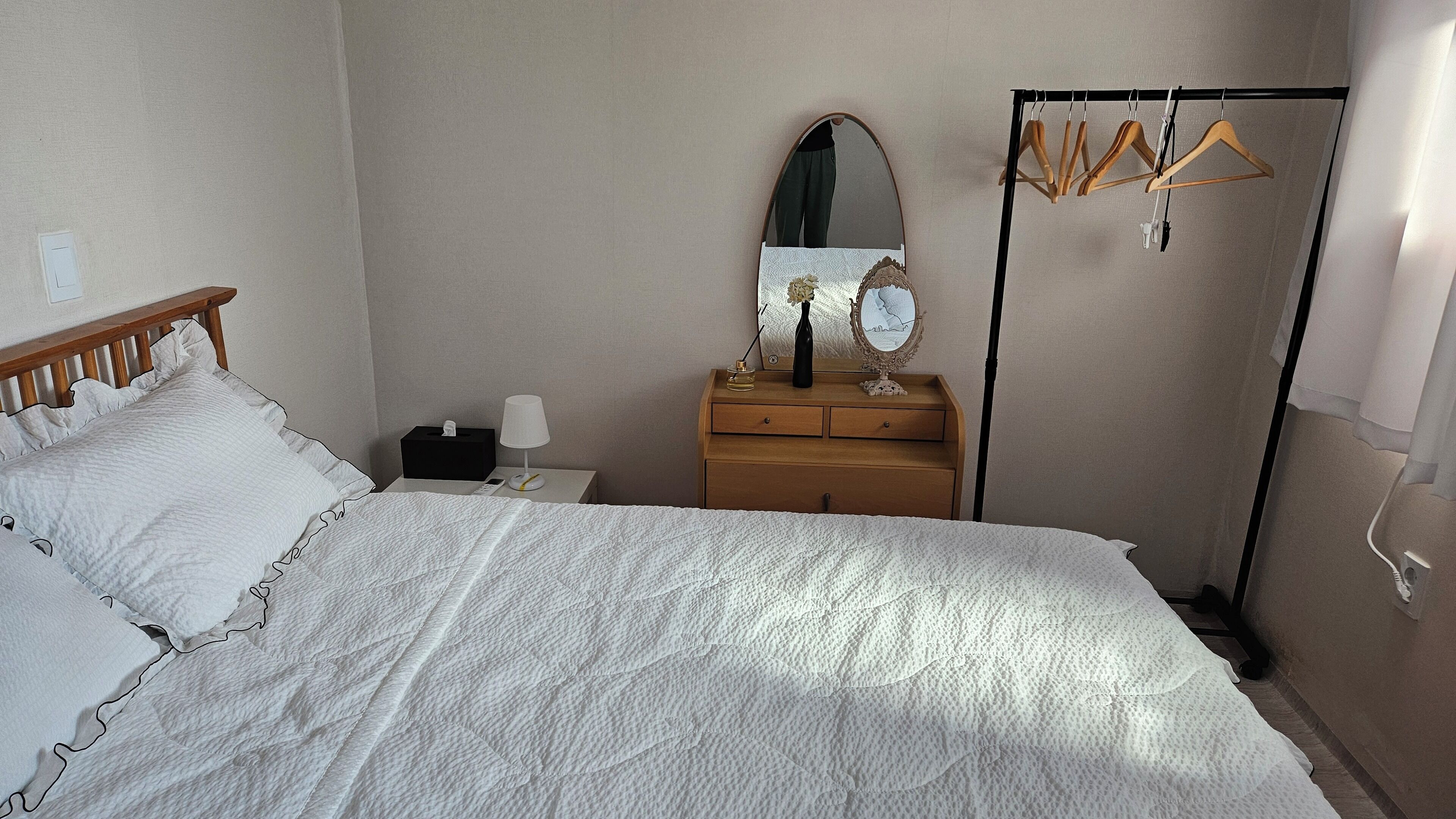 A neatly made bed with a white quilt, a wooden headboard, a bedside table with a lamp, a mirror, and a wooden dresser with a mirror.