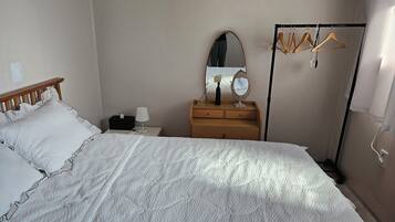 A neatly made bed with a white quilt, a wooden headboard, a bedside table with a lamp, a mirror, and a wooden dresser with a mirror.