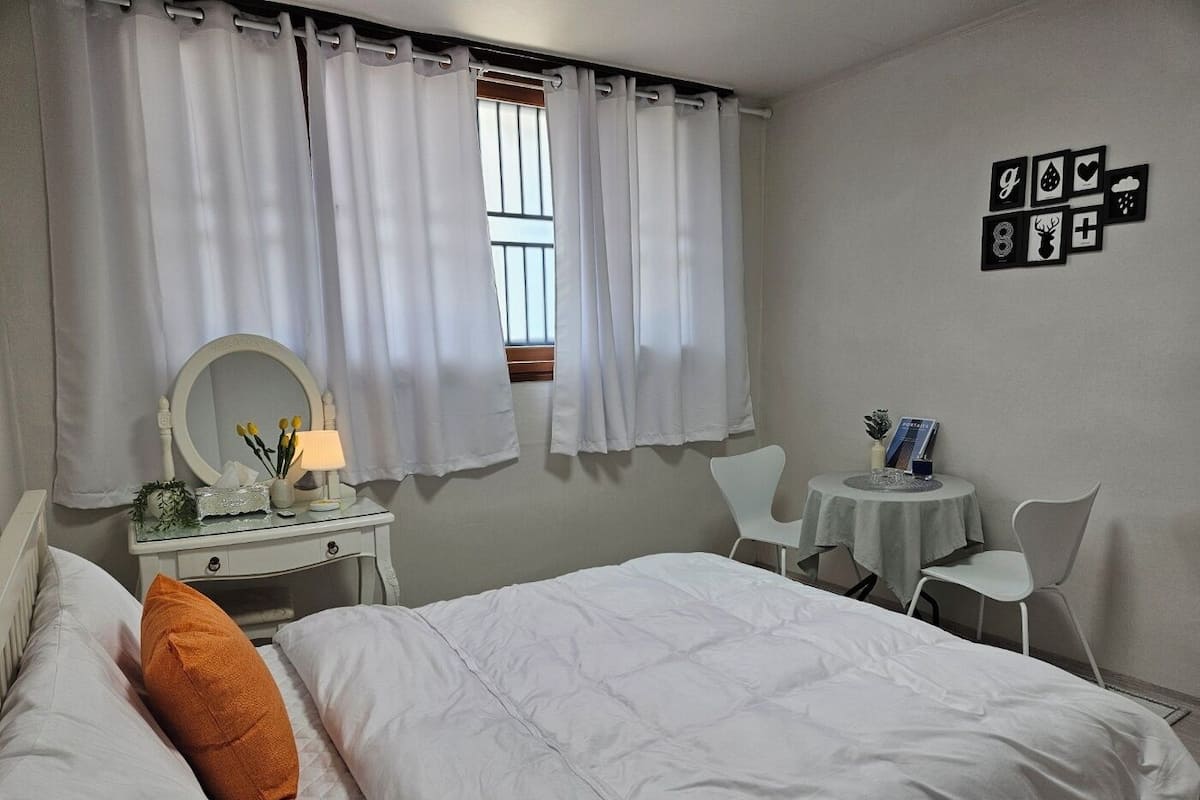 Un dormitorio con cama, una mesita de noche con lámpara, un espejo y una pequeña mesa con un libro.