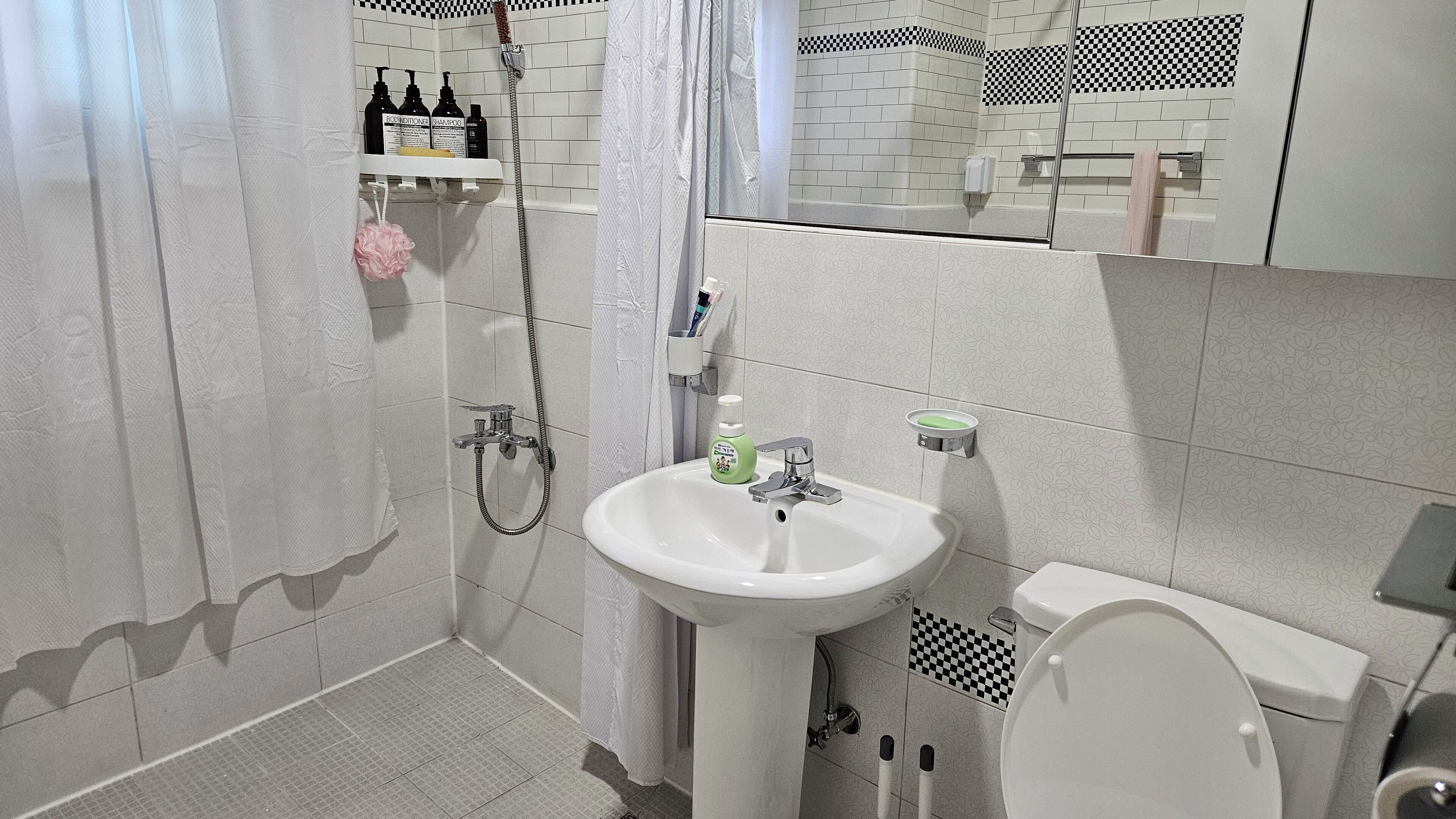 A bathroom with a white sink, toilet, and shower. There are toiletries on a shelf and a towel hanging on the wall.