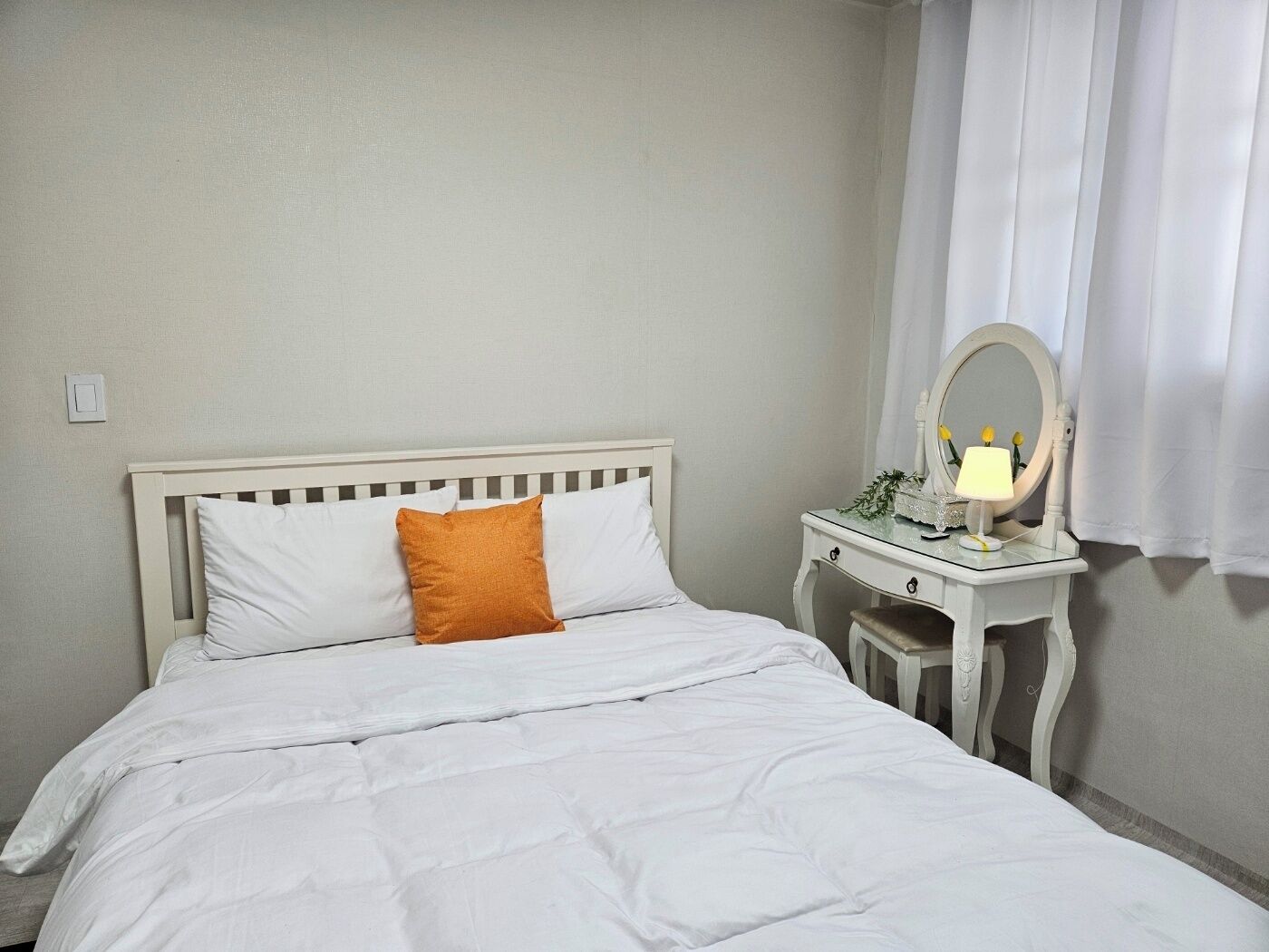 A neatly made bed with white bedding and an orange pillow, a white bedside table with a mirror and a lamp, and a white curtain partially drawn to the side.