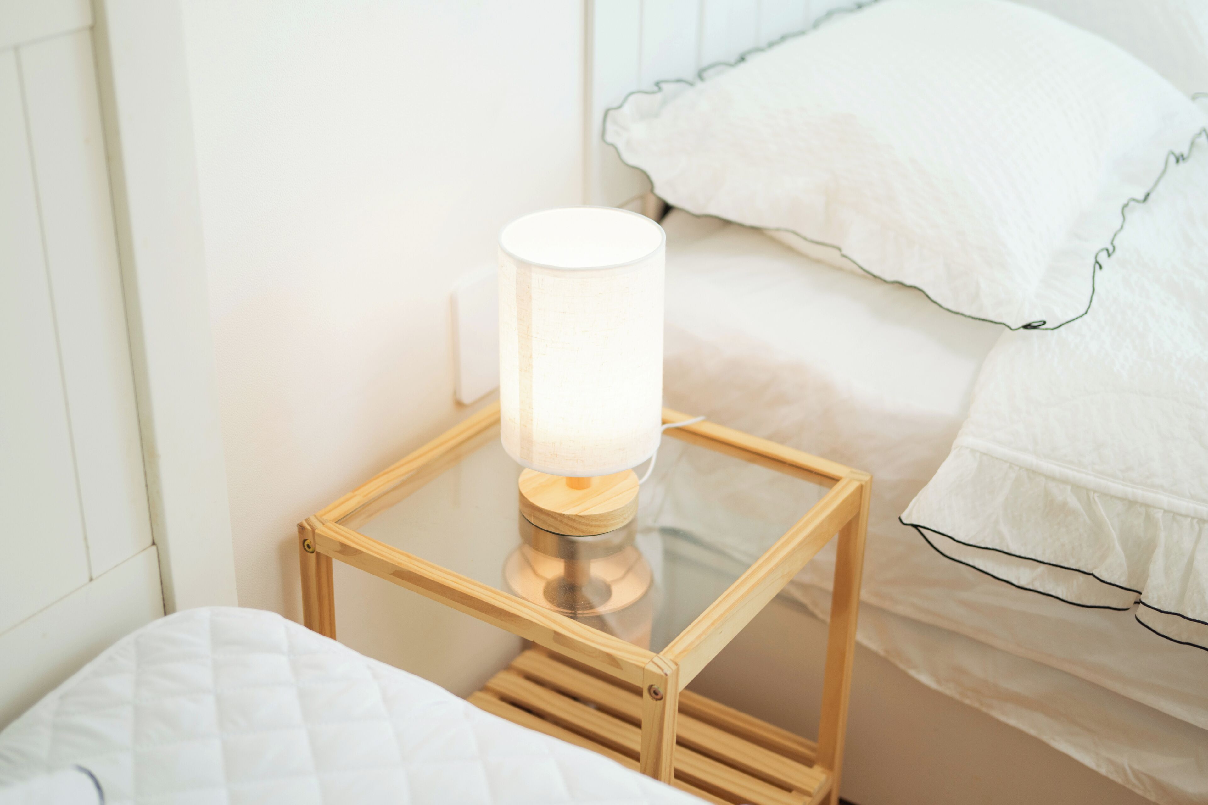 Una mesita de noche con una lámpara, una cama con ropa blanca y una pared blanca.