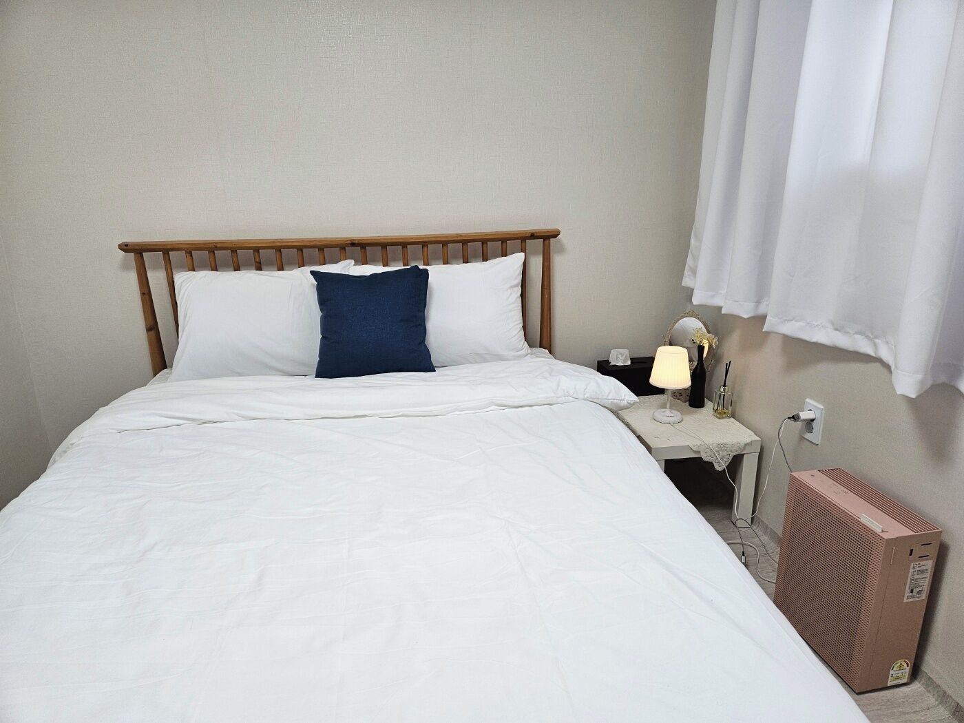 A neatly made bed with white and blue pillows, a bedside table with a lamp, a mirror, and a heater.