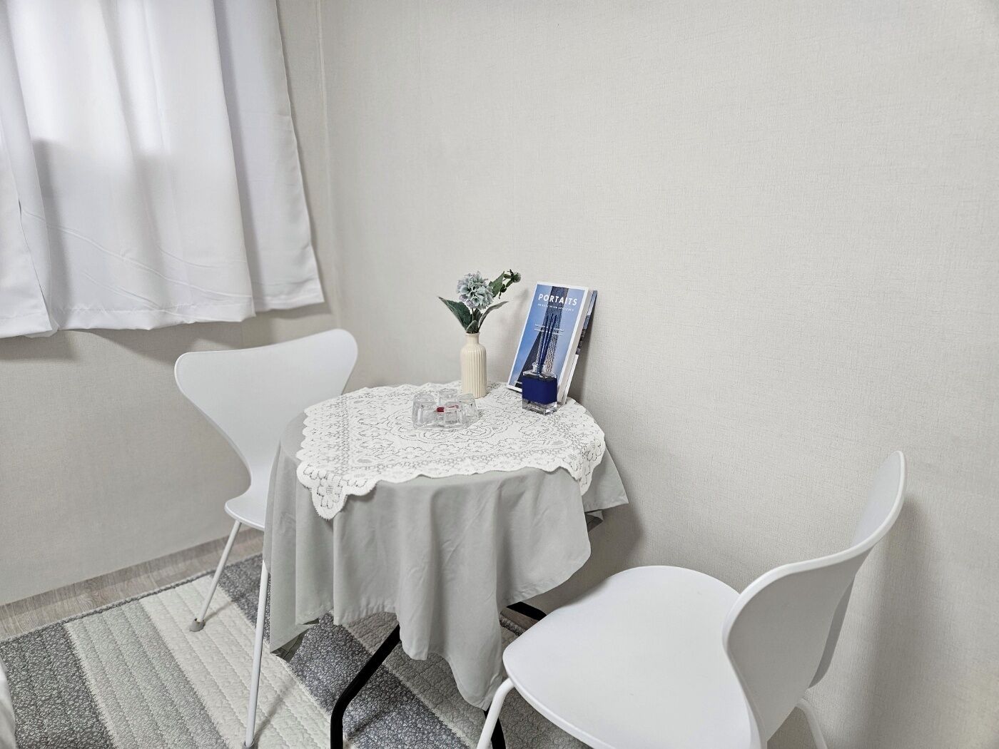 A small, neatly arranged room with a round table covered by a white tablecloth, two white chairs, a lace table runner, a vase with flowers, and a book titled "PORTAITS".