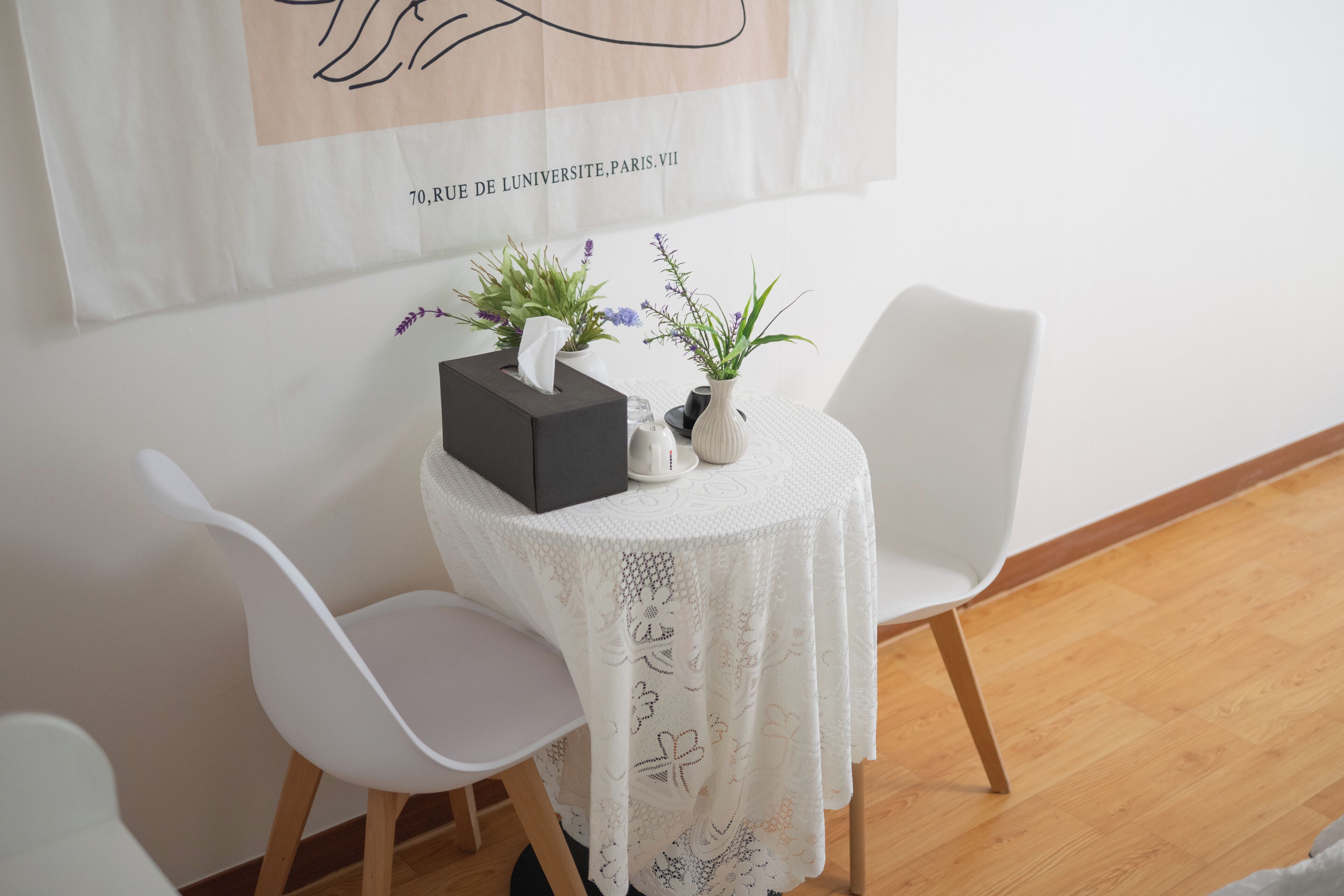 Una mesa pequeña con mantel de encaje blanco, dos sillas blancas y un florero con plantas.