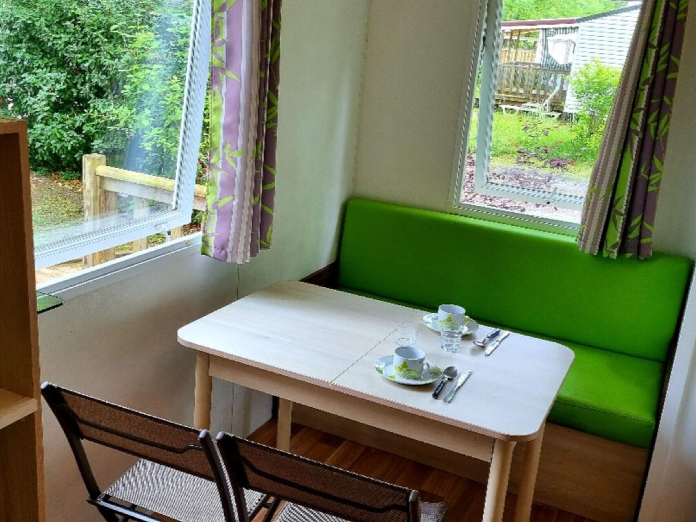 Une petite pièce avec une table et un banc vert. Il y a une fenêtre avec des rideaux, et de là, on peut voir un jardin et une maison.