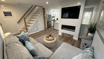 A modern living room with a sofa set, a fireplace, a television, and a kitchen in the background.