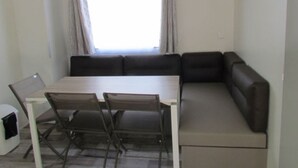 A compact living space with a dining table and chairs, a sofa, and a window with curtains. - 31m² Mobile Home with Terrace - 3 Bedrooms, Sleeps 6 (La Grande-Motte)