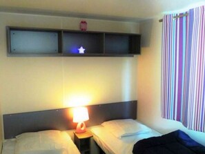 Quarto de hotel com duas camas, mesa de cabeceira com abajur, prateleira sobre as camas e cortina listrada.