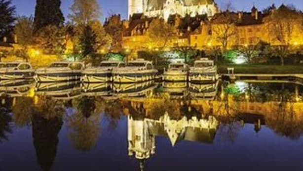 Un edificio histórico con una torre que se refleja en un cuerpo de agua tranquilo, rodeado de árboles y barcos.