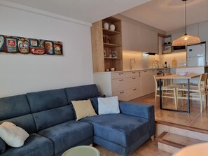A modern living room with a blue sofa, a dining area with wooden chairs, a kitchen with light wood cabinets, and a hanging light fixture. - Modern apartment with terrace and pool 50m from the beach (L'Escala)