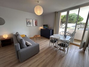 A modern living room with a grey sofa, a small round table, a TV on a cabinet, and a dining area with white chairs. - Studio with Cabins - Sleeps 4 - Parking - Close to Beach (Saint-Hilaire-de-Riez)