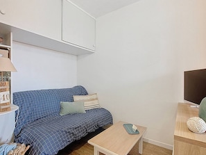 Ein kleiner, einfach eingerichteter Raum mit einem blauen Sofa, einem kleinen Holztisch und einem Fernseher auf einem Holzgestell.