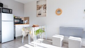 Cozinha moderna com geladeira, micro-ondas e cafeteira. Há uma mesa de jantar com cadeiras e um banquinho pequeno. Um espelho decorativo está pendurado na parede.