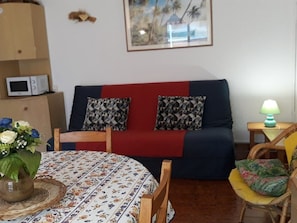 A living room with a sofa, a table with a floral tablecloth, a chair, a lamp, and a framed picture on the wall. - 2-Rooms Apartment, Bormes-les-Mimosas, Beach (Bormes-les-Mimosas)