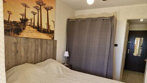 A bedroom with a large bed, a headboard, a nightstand with a lamp, a dark curtain, and a door. - 2 Bedroom Apartment, Air Conditioning, Parking, Sleeps 4 (Bormes-les-Mimosas)
