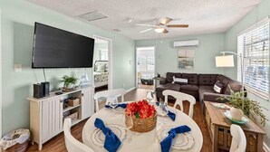 A living room with a TV, a ceiling fan, a dining table set with glasses and a centerpiece, and a brown sofa. - Sunny Isles Beach Cottage, Indian Shores! (Indian Shores)