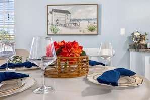 Una mesa de comedor dispuesta con cristalería transparente, una cesta de mimbre con flores y un cuadro en la pared.