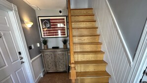 A wooden staircase with a handrail, a wall-mounted American flag, a cabinet with two potted plants, and a light fixture. - Spacious Three Room Suite Downtown Newport #16 1 Bedroom Home (Newport)