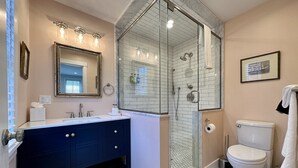 Un baño con ducha envuelta en vidrio, un inodoro blanco, un tocador azul oscuro y un espejo con luces arriba.