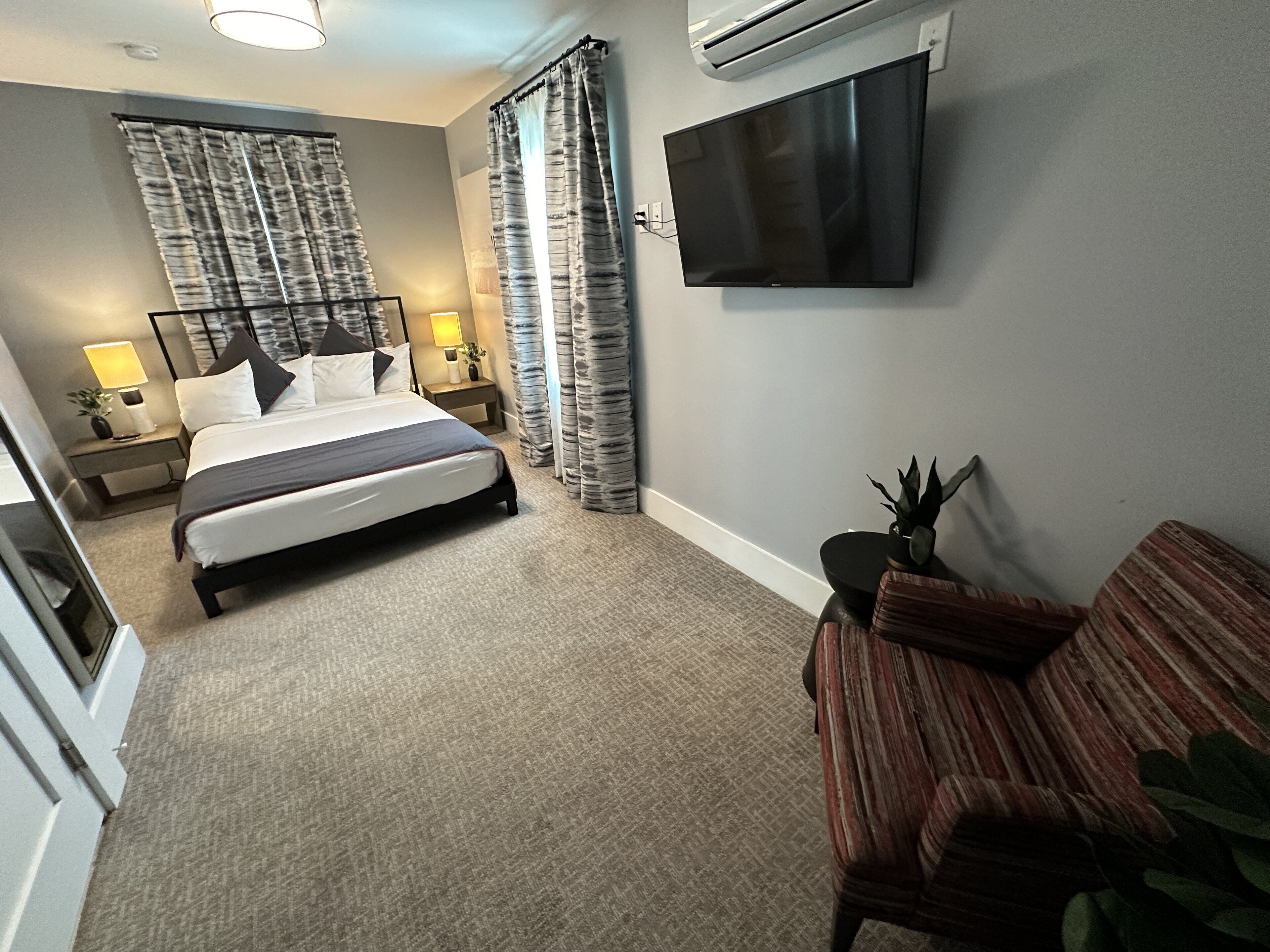 A hotel room with a bed, a TV mounted on the wall, a desk with a lamp, and a chair.