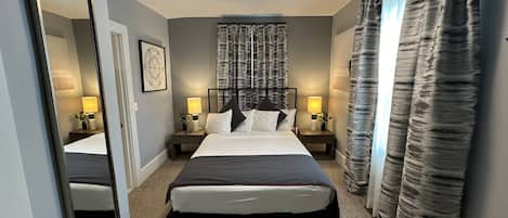 A hotel room with a large bed, two bedside tables with lamps, a ceiling light, a large mirror, and a window with curtains.